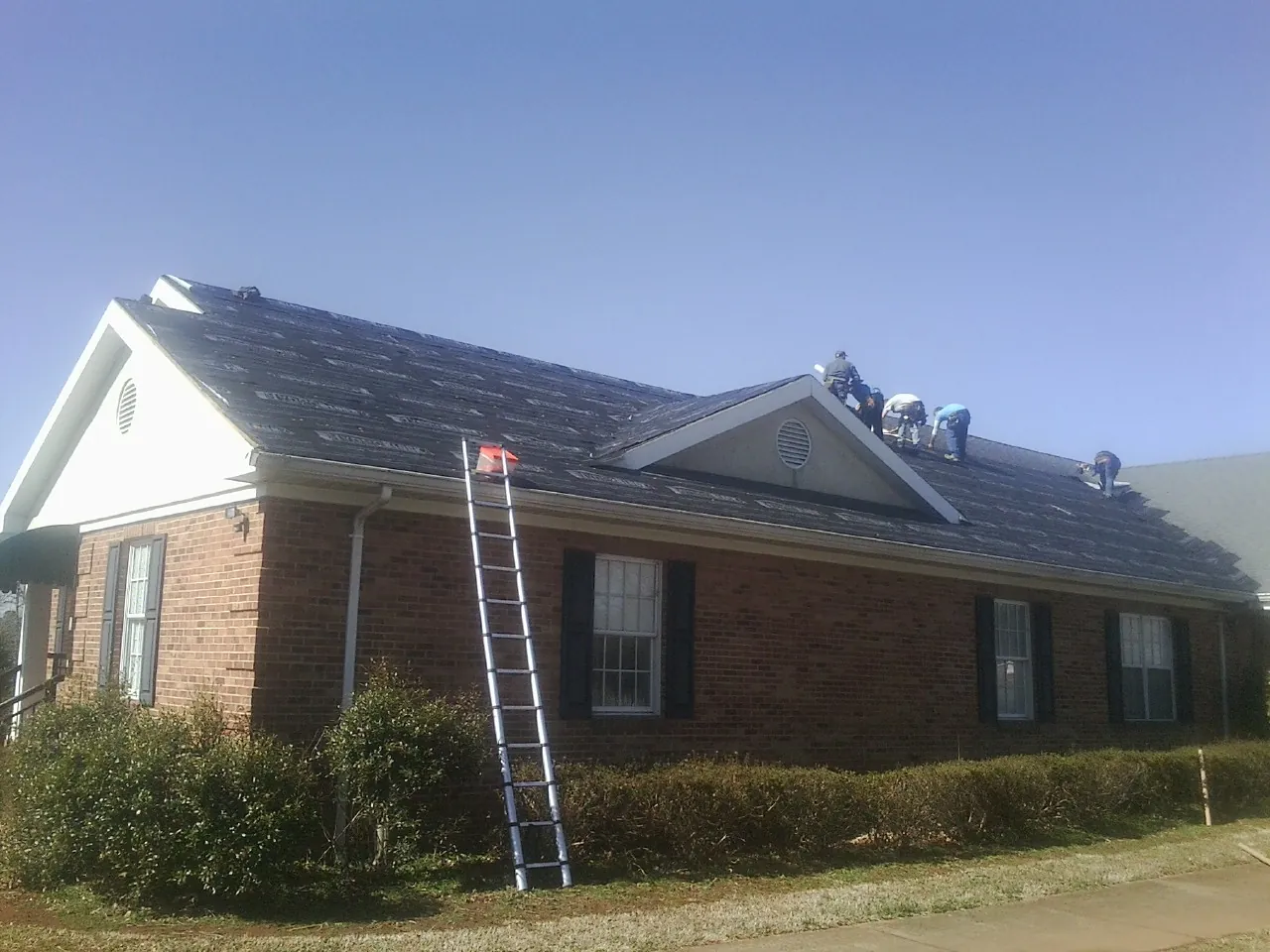 Crew tearing shingles off a brick ranch roof with a ladder set — Metal Roof Repair work by our crew in Port Haywood