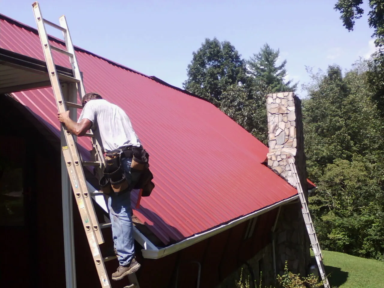 Roofer on a ladder beside a red metal roof and stone chimney — Soffit & Fascia Repair by Roofing Brothers in Port Haywood